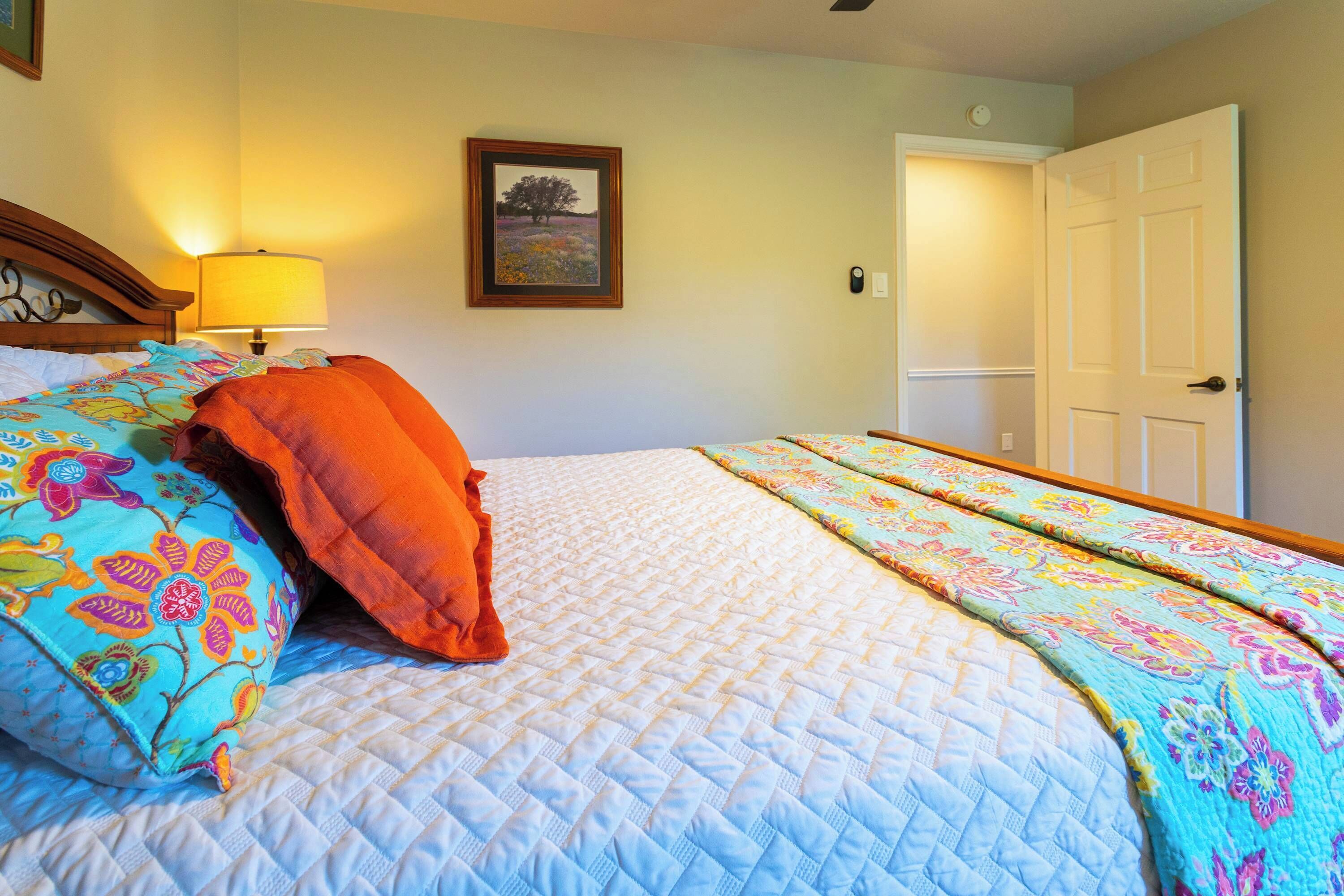 Una cama bien tendida con ropa de cama floral y cubrecama acolchado, una lámpara de noche y un cuadro en la pared.