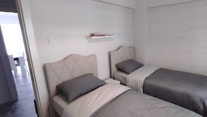 Une chambre avec deux lits simples, une petite étagère et une porte donnant sur une autre pièce.