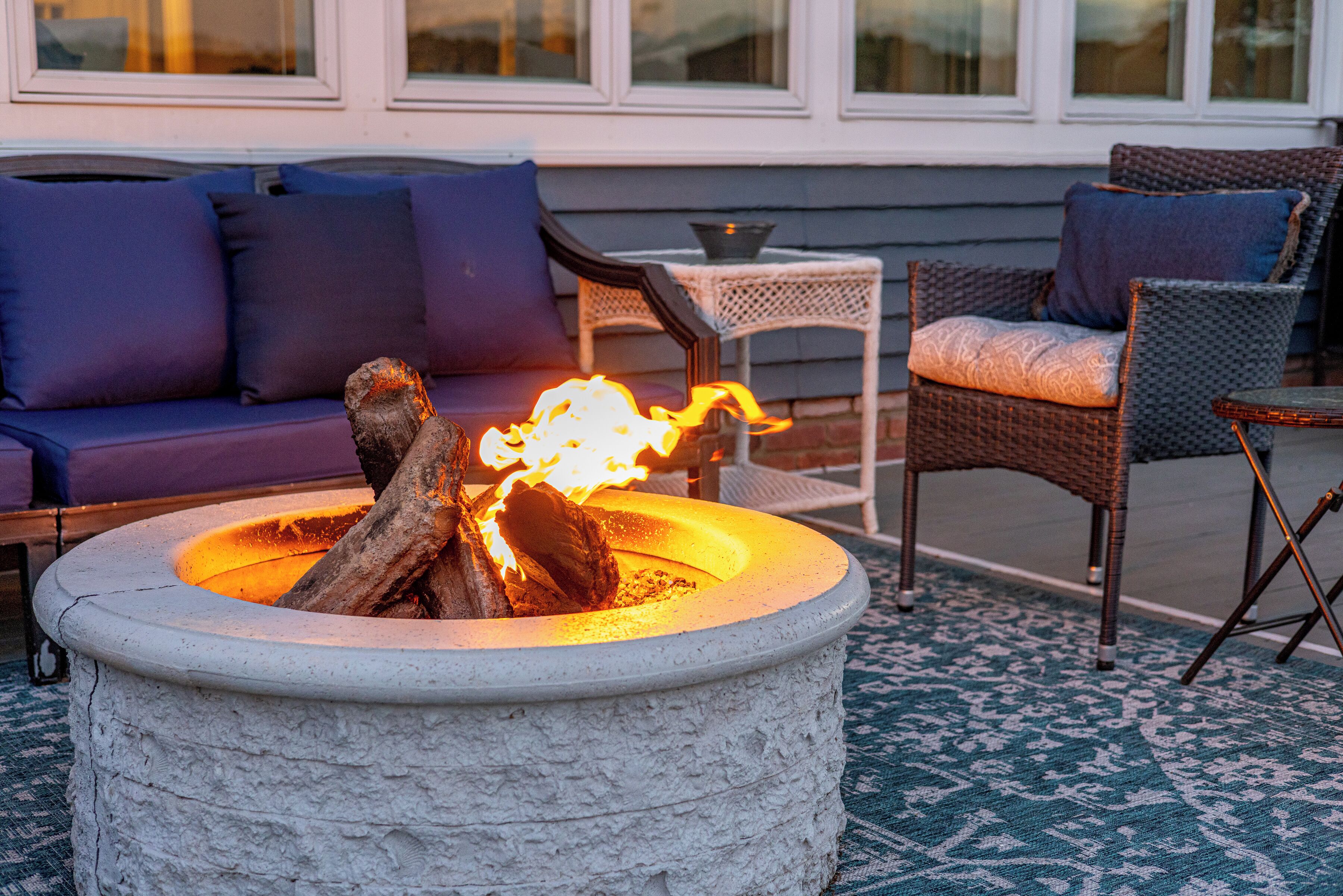 A stone fire pit with logs, a sofa with blue cushions, and wicker chairs.