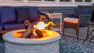 A stone fire pit with logs, a sofa with blue cushions, and wicker chairs.