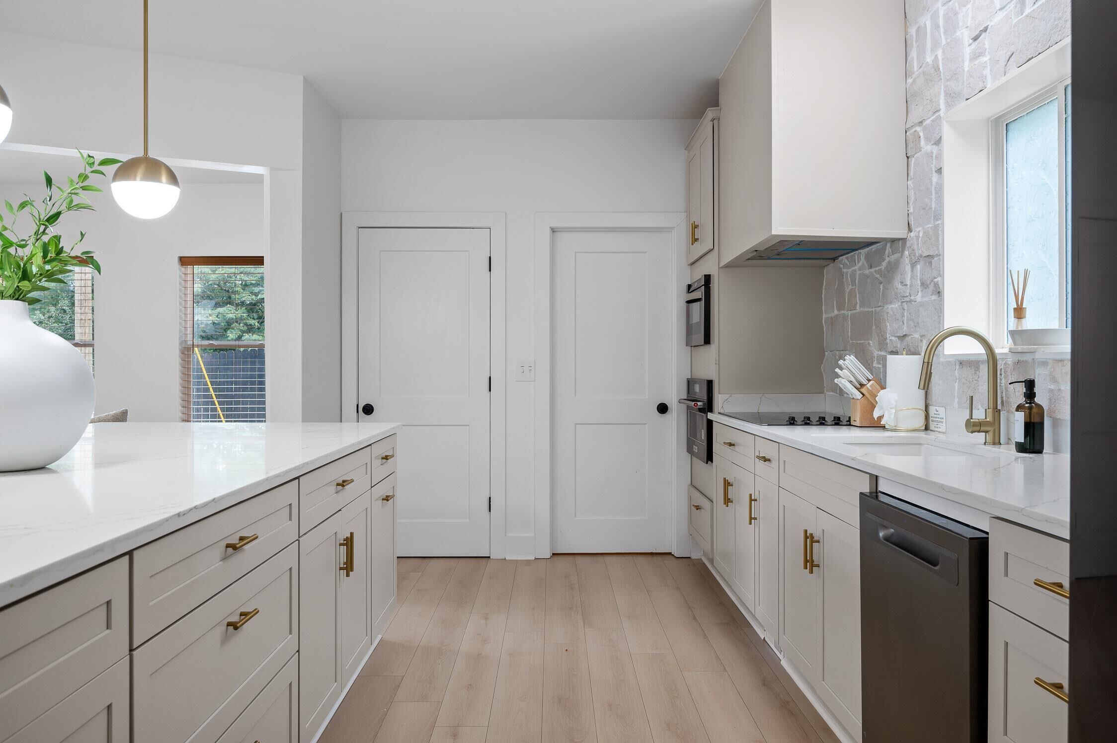 Una cocina moderna con muebles blancos, lavavajillas, estufa y lavabo. Hay una ventana con vistas a árboles y una puerta que da a otra habitación.