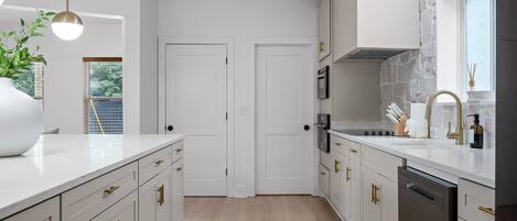 Una cocina moderna con muebles blancos, lavavajillas, estufa y lavabo. Hay una ventana con vistas a árboles y una puerta que da a otra habitación.
