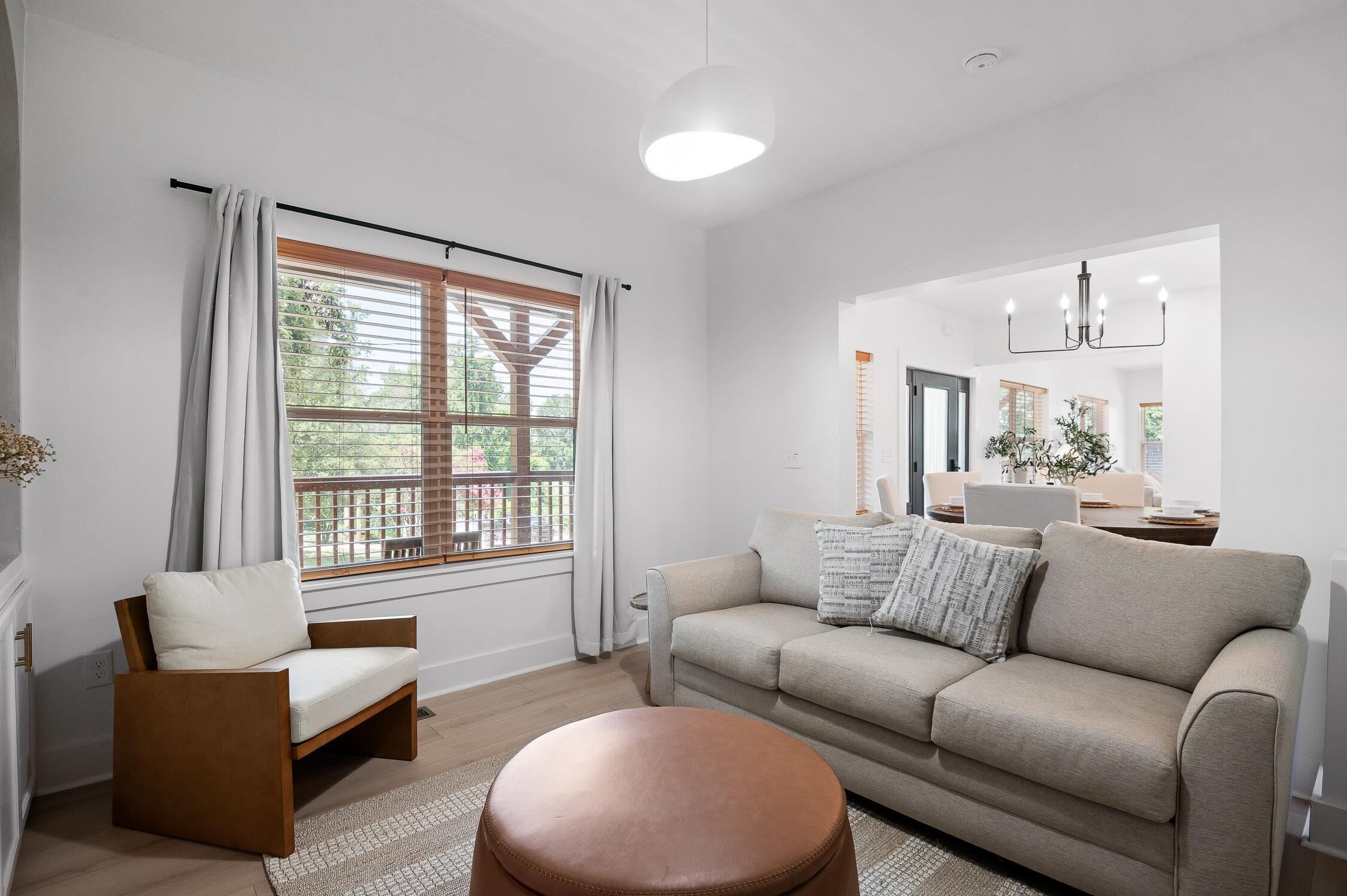 A modern living room with a sofa, a chair, a round ottoman, and a large window with blinds.