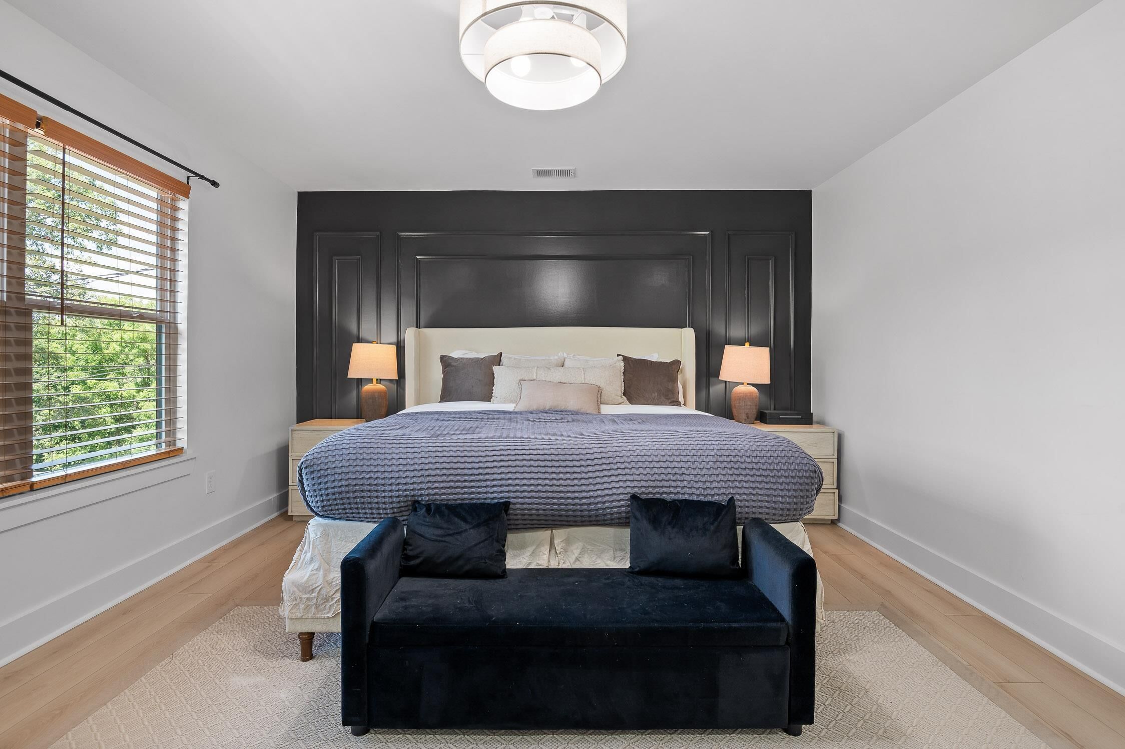 A modern bedroom with a large bed, two bedside lamps, a dark headboard, and a bench.