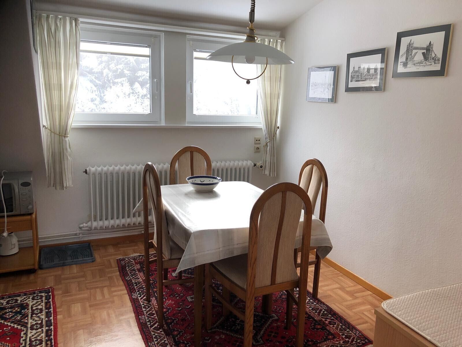 A dining area with a table, chairs, a bowl, a radiator, a window with curtains, and framed pictures on the wall.