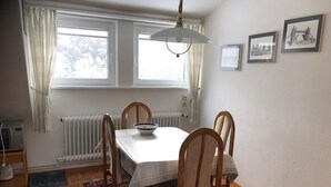 Un coin repas comprenant une table, des chaises, un bol, un radiateur, une fenêtre avec des rideaux et des tableaux encadrés accrochés au mur.