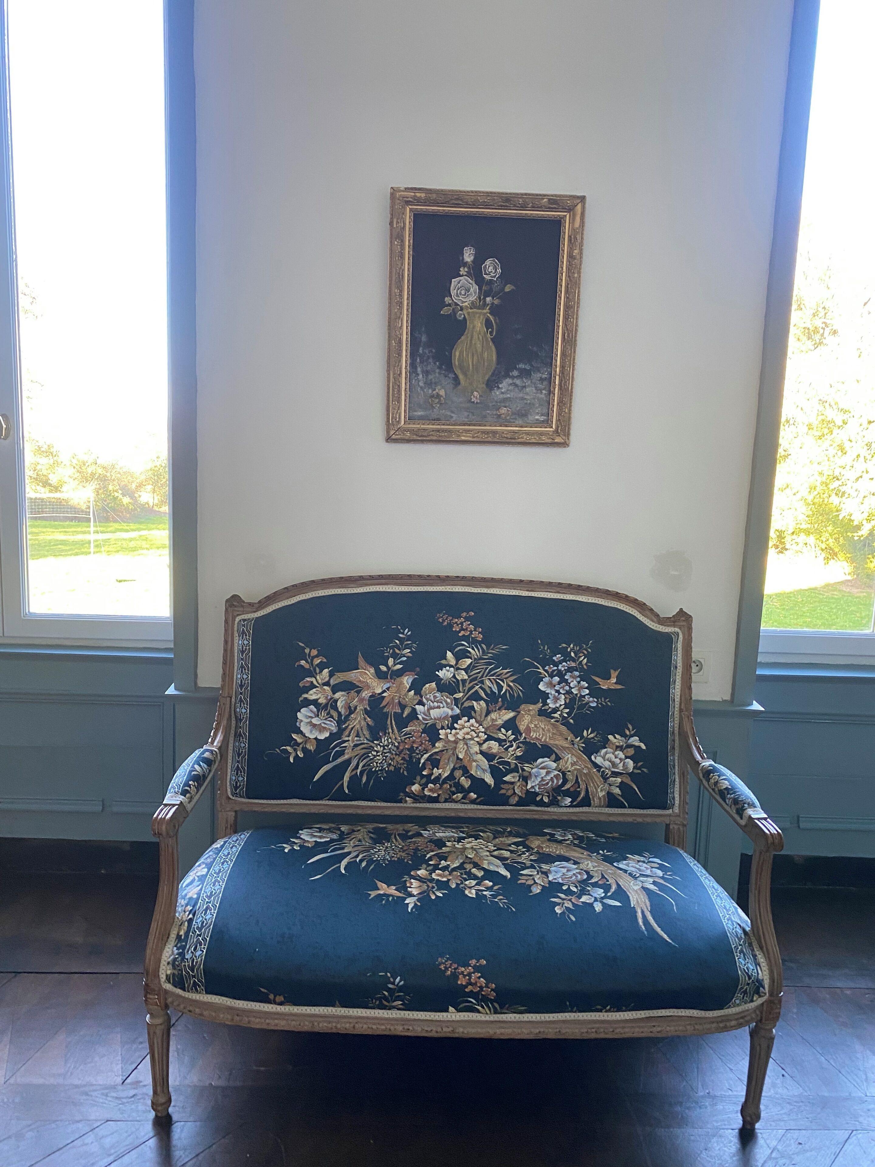 En ældre blå polstret stol med detaljerede blomstermønstre, placeret i et rum med store vinduer og et indrammet maleri af en vase med blomster på væggen.