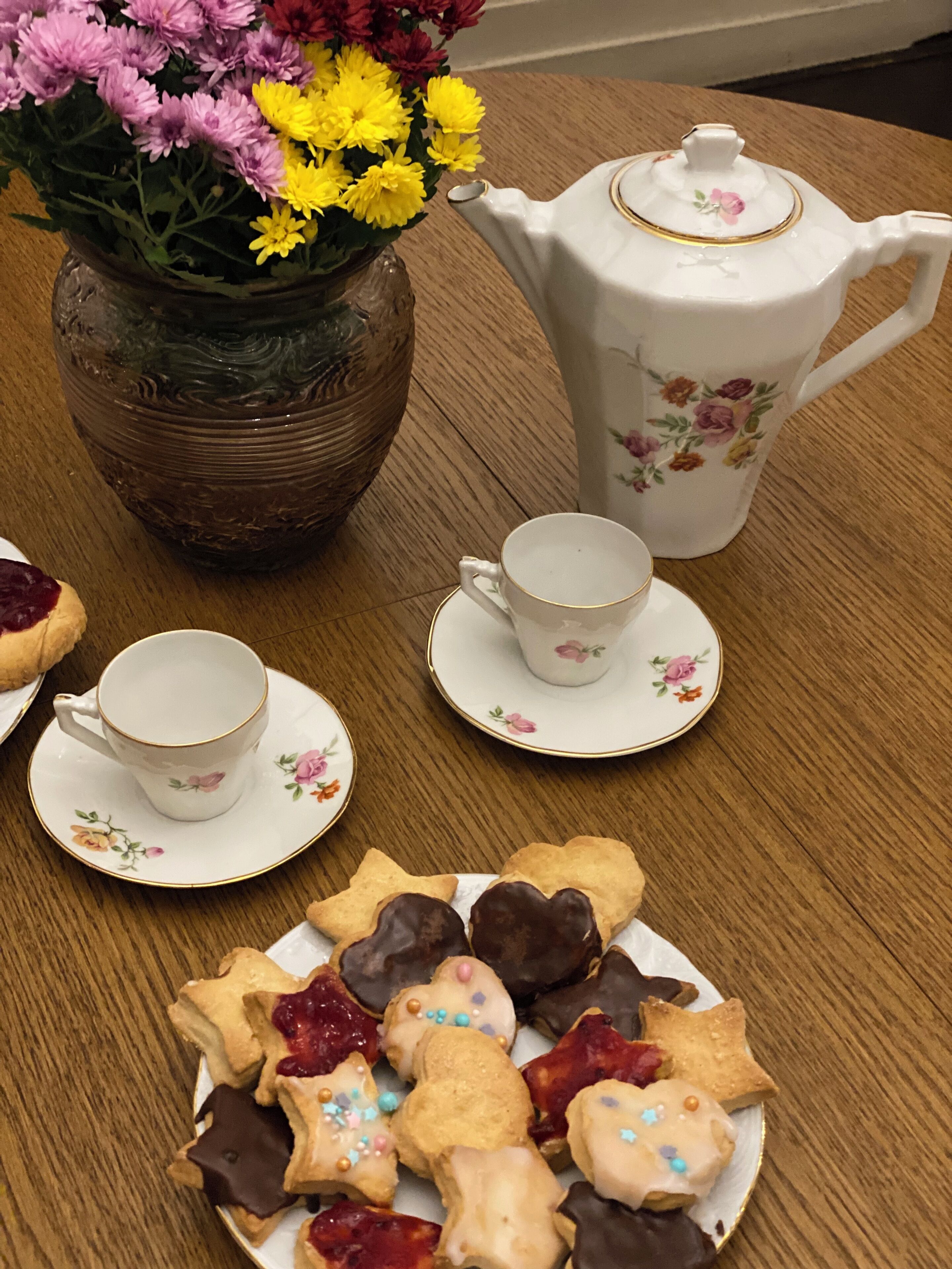 Uma mesa de madeira com uma chaleira floral, duas chávenas, pires e um prato de biscoitos decorados.