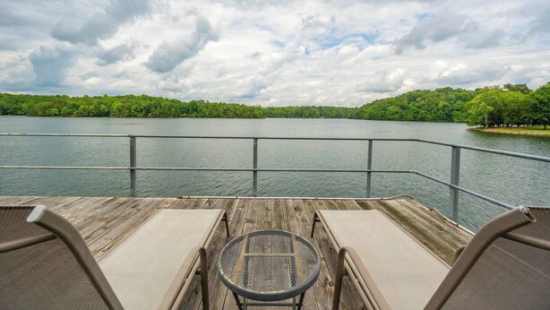 A wooden deck with two chairs and a table, overlooking a calm lake with a forested shoreline.