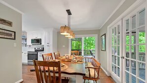 A dining area with a wooden table and chairs, a hanging light fixture, and a view of the outdoors through large windows.