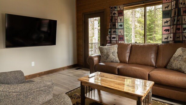 A living room with a brown leather sofa, a wooden coffee table, a grey armchair, a flat-screen TV, and a window with a view of trees.