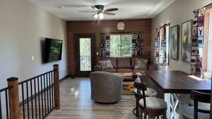 A spacious living room with a brown leather sofa, a round coffee table, and a wooden dining table with two chairs. A ceiling fan and a clock are visible on the wall.