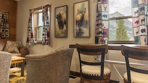 A cozy room with a wooden floor, a brown leather sofa, patterned chairs, and a window with checkered curtains featuring animal prints.