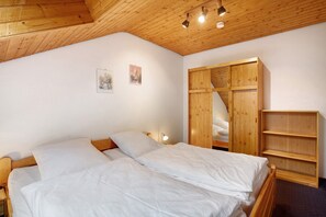 Een kamer met houten plafond, een bed met wit beddengoed, een houten garderobe en een klein wandrek.