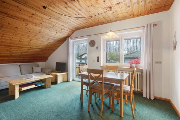 A cozy room with a sloped ceiling, a wooden dining table with chairs, a sofa, a small TV, and a window with curtains.
