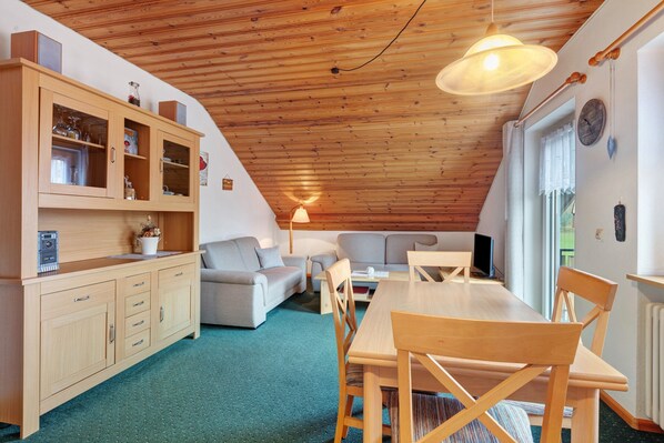 A cozy attic room with a wooden ceiling, a dining table with chairs, a sofa, a cabinet with glass doors, and a lamp.
