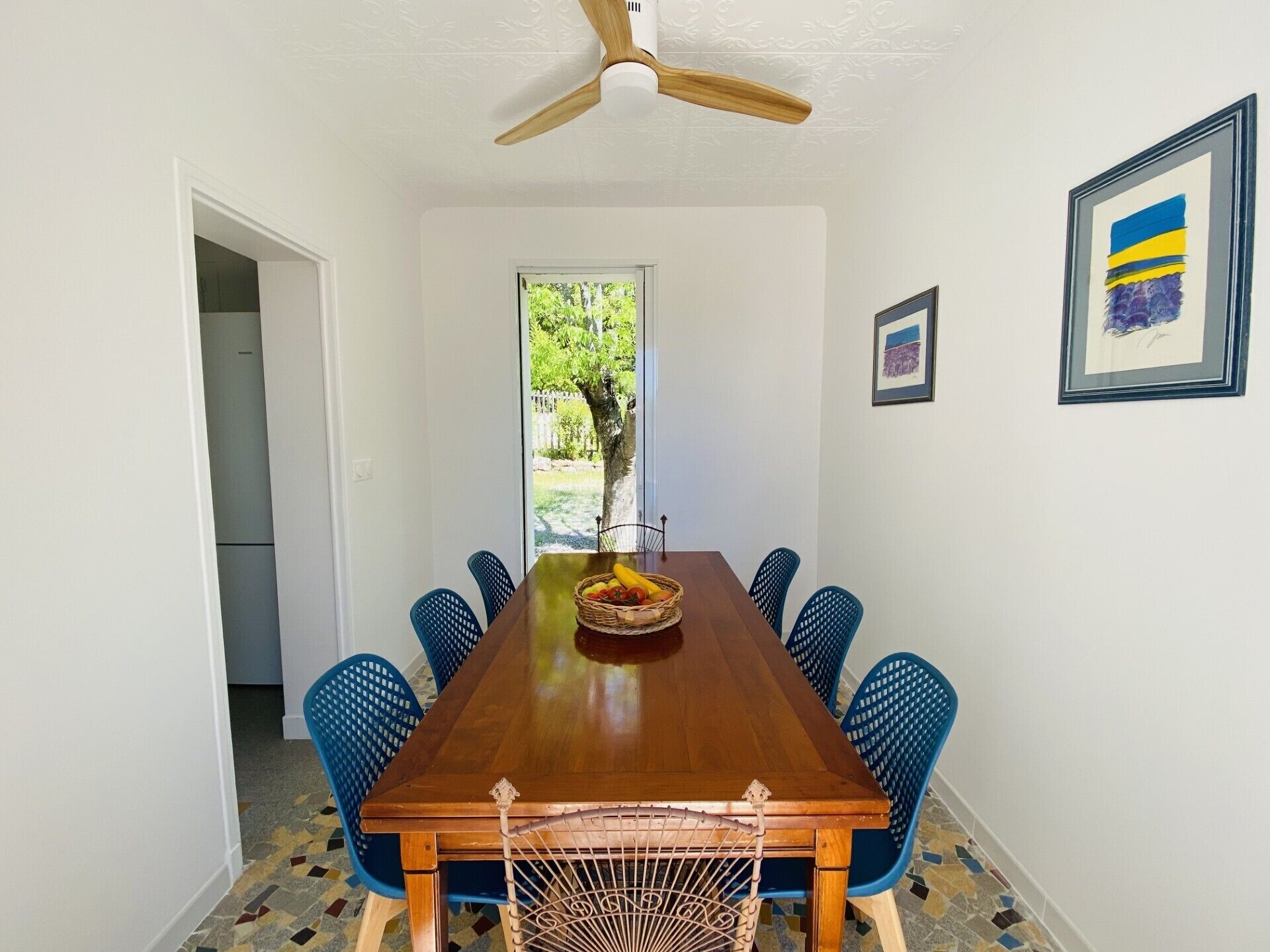 Comedor con mesa de madera, sillas azules, ventilador de techo y ventana con vista a follaje.