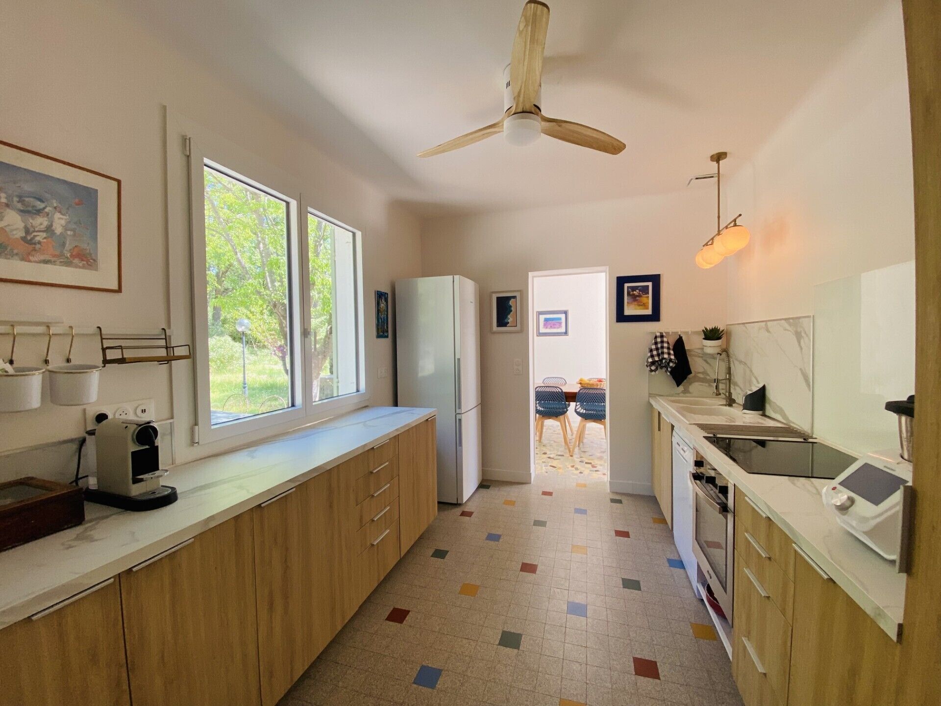 Una cocina compacta con armarios de madera, un refrigerador blanco, un ventilador de techo y vistas al jardín a través de ventanas amplias.