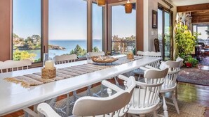 A dining area with a long table, white chairs, and a view of the sea.