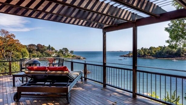 A wooden deck with a lounge chair and umbrella, overlooking a body of water and distant land.