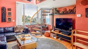A living room with a black leather sofa, a glass coffee table, a TV, and a staircase leading to a balcony with a view.
