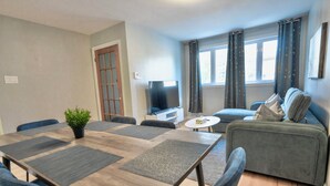Una sala de estar moderna con una mesa de comedor de madera, sillas azules, un sofá gris, un televisor y una ventana con cortinas.
