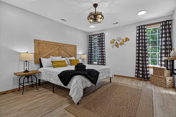 A modern bedroom with a wooden headboard, a bed with white bedding and a black throw, two bedside tables with lamps, a large window with checkered curtains, and a decorative wall piece.