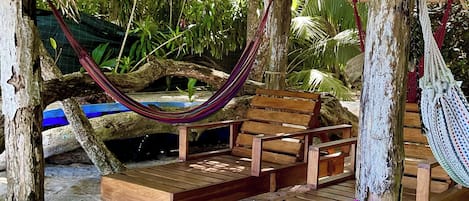 A wooden bench and hammock under a thatched roof, surrounded by tropical plants.