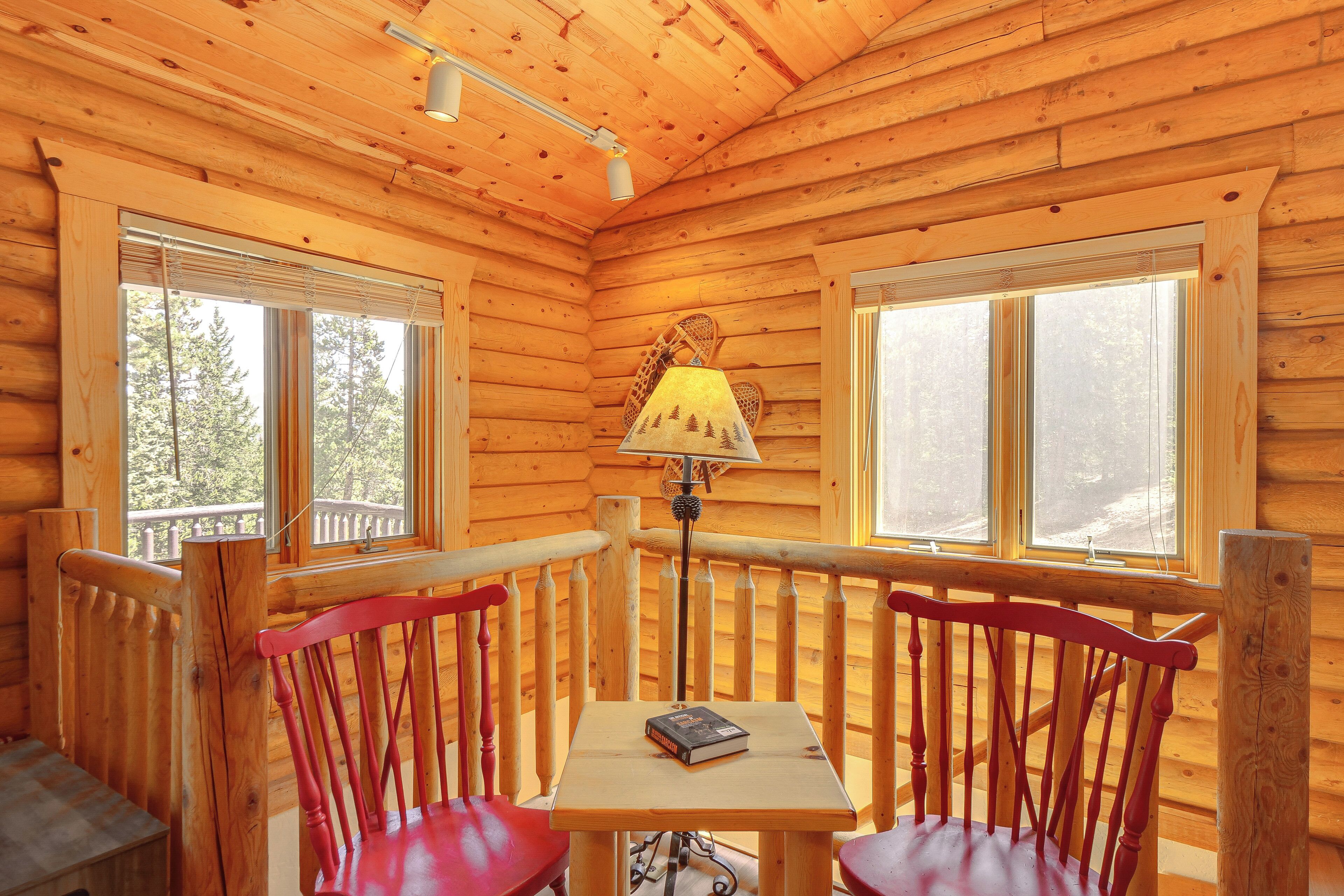 A wooden cabin interior with a table and chairs, a lamp, and large windows.