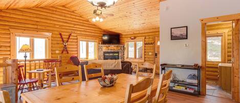 A wooden cabin interior with a dining table, chairs, a fireplace, a television, and a stone wall.