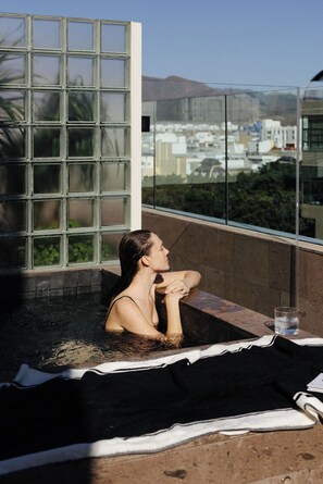Une personne se détend dans un bain à remous, avec vue sur des bâtiments et des montagnes, grâce à une grande fenêtre.