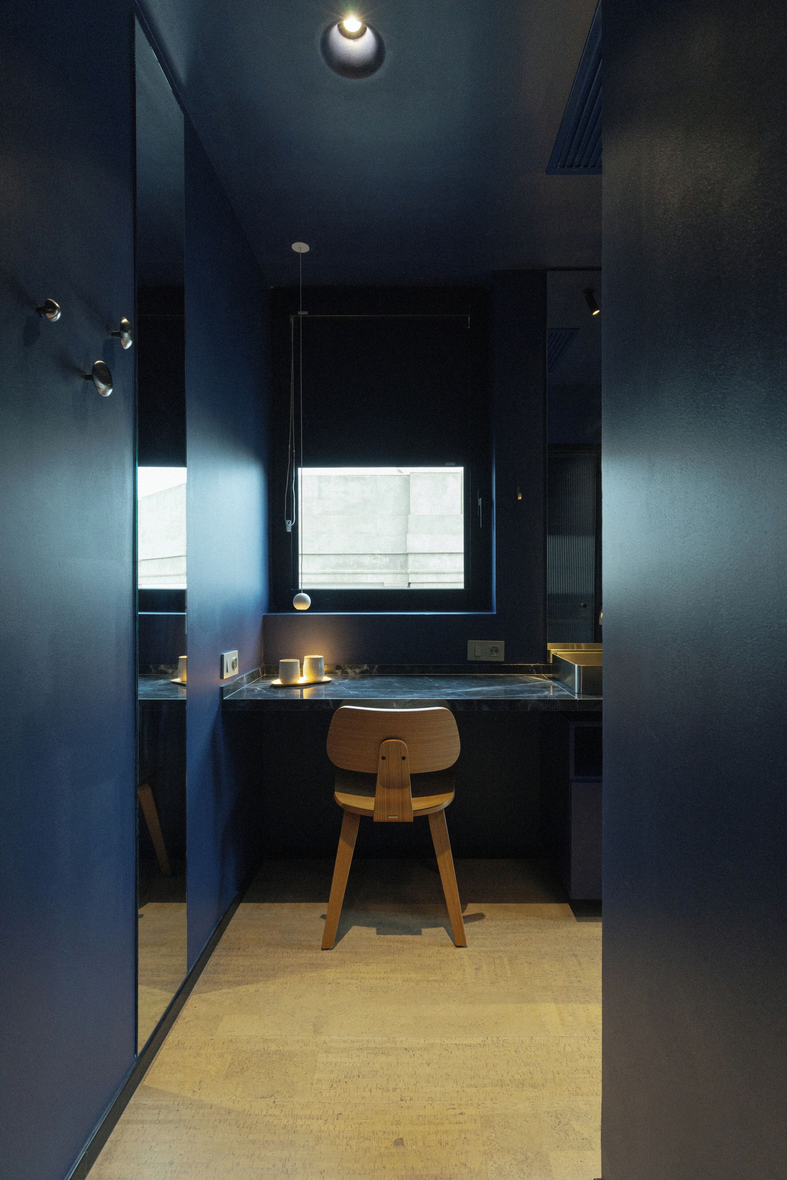 Een moderne badkamer met een donkerblauwe kleurstelling, een houten stoel en een raam dat natuurlijk licht binnenlaat.