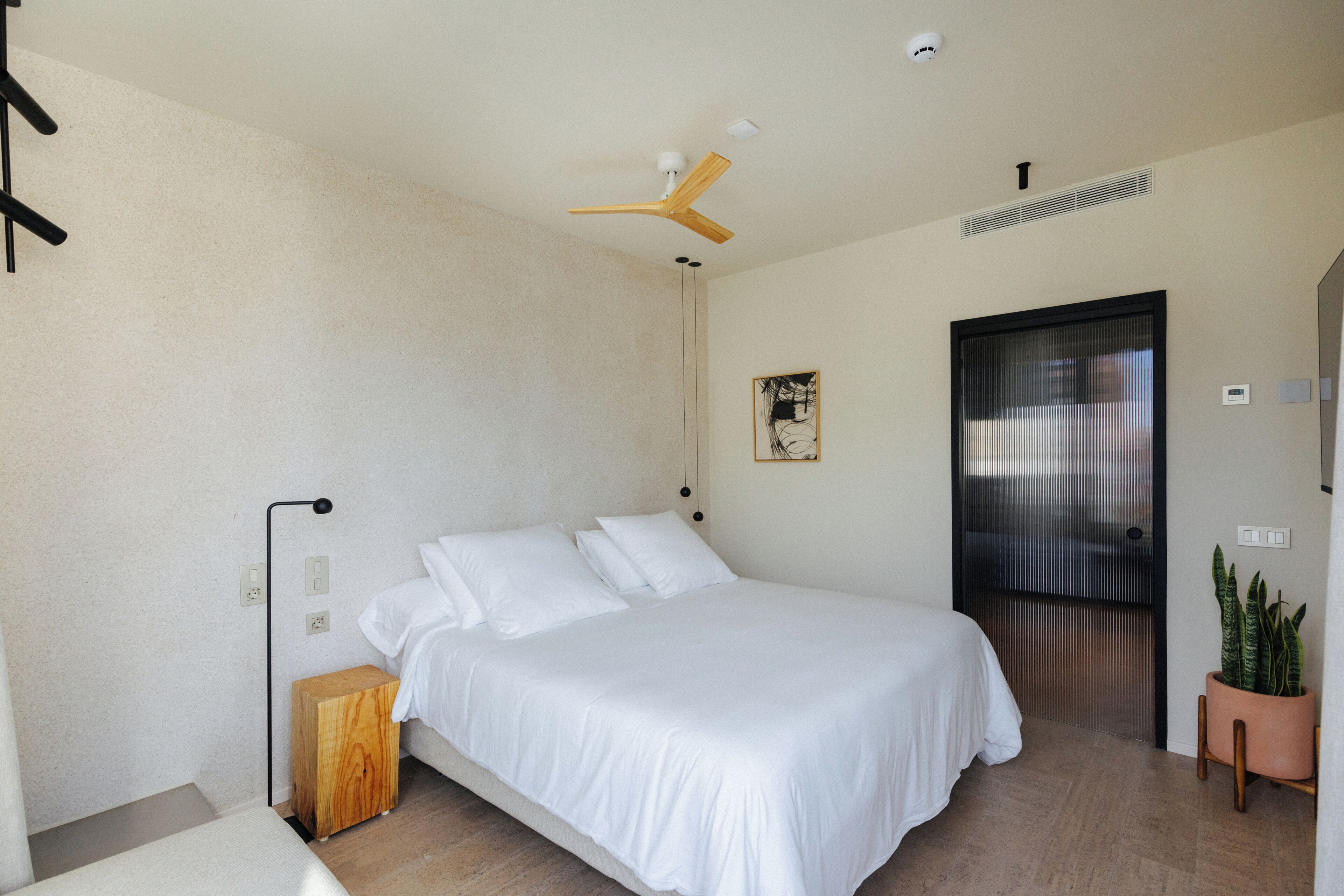 A modern bedroom with a bed, a ceiling fan, a small wooden nightstand, a potted plant, and a door with a view.