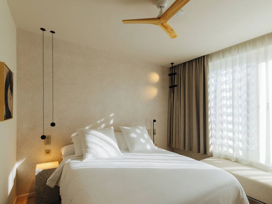A hotel room with a bed, a ceiling fan, a window with curtains, and a wall-mounted light fixture.