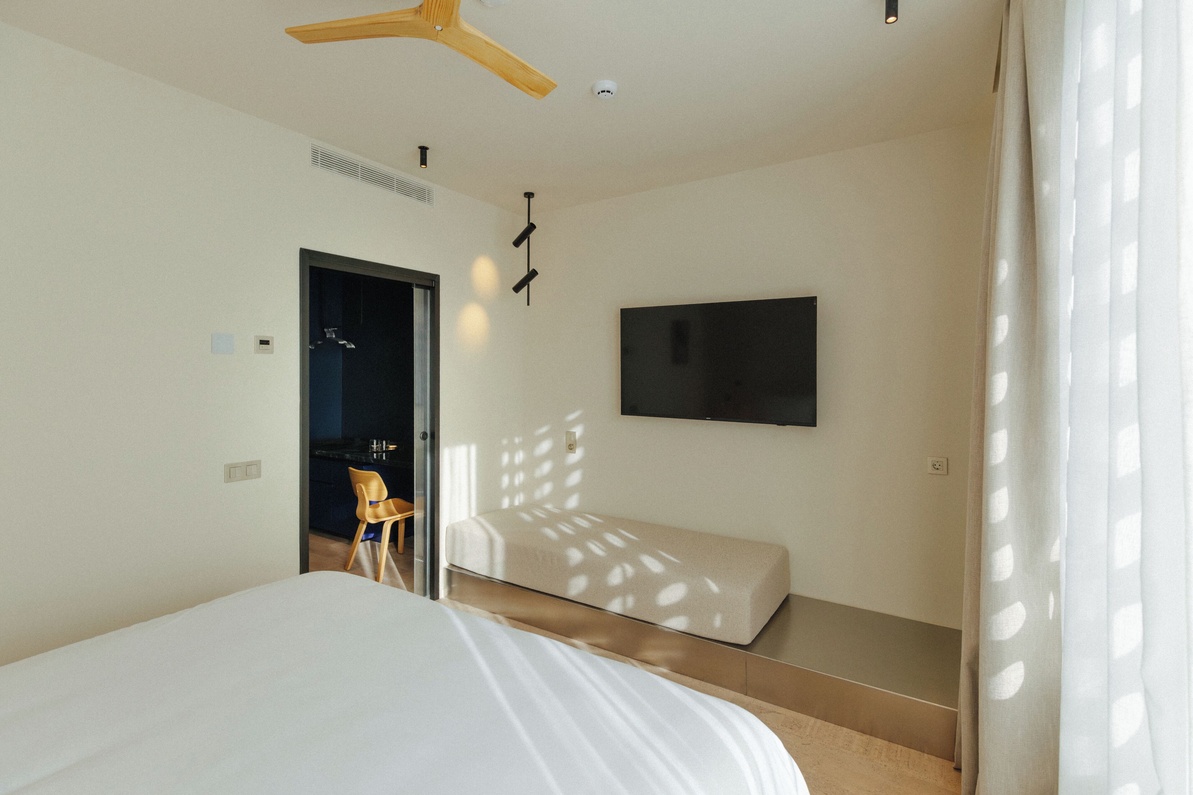A modern hotel room with a bed, a TV mounted on the wall, a ceiling fan, and a door leading to a bathroom.