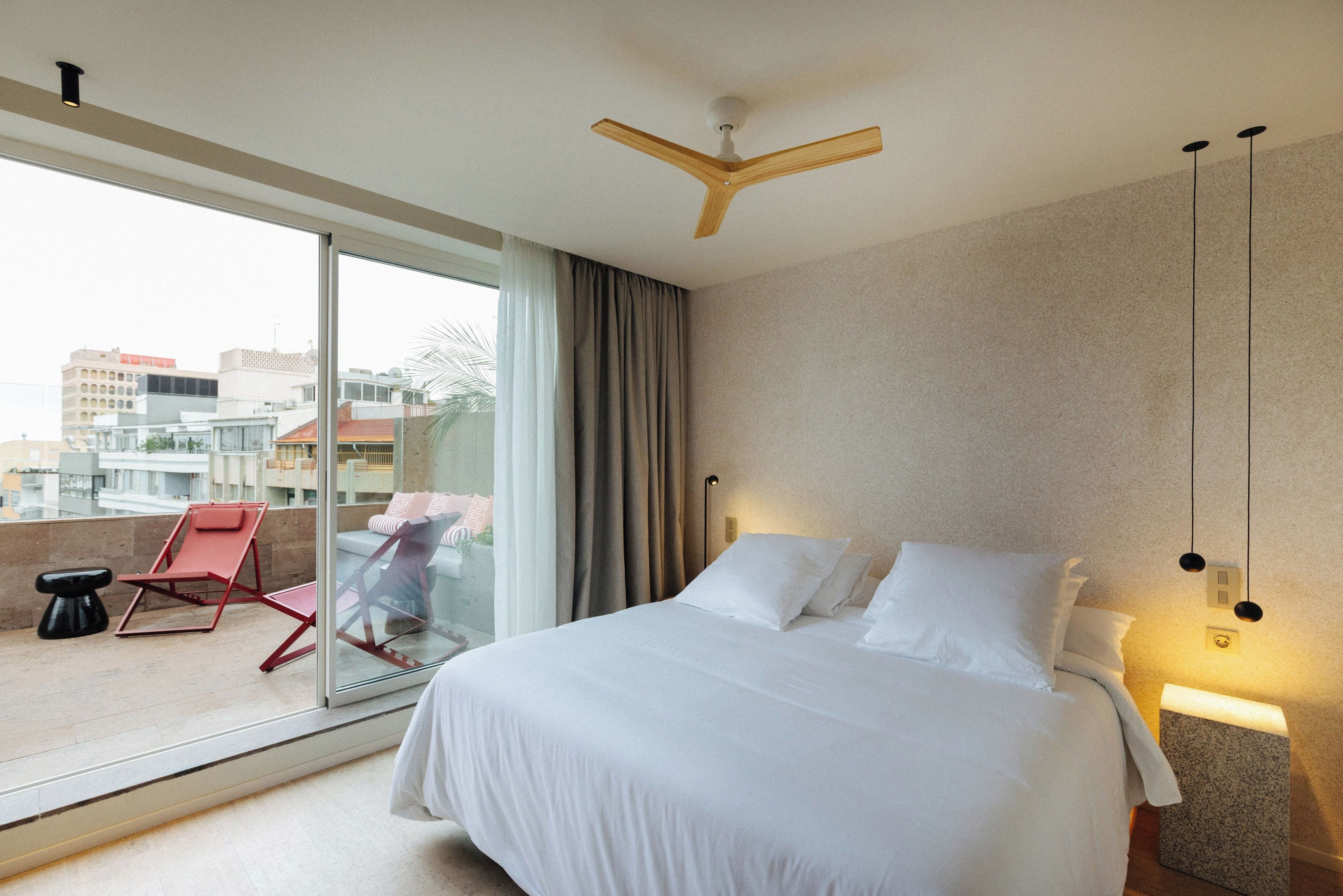 Une chambre d’hôtel avec un grand lit, un ventilateur de plafond, un balcon avec une chaise rouge et une vue sur la ville.