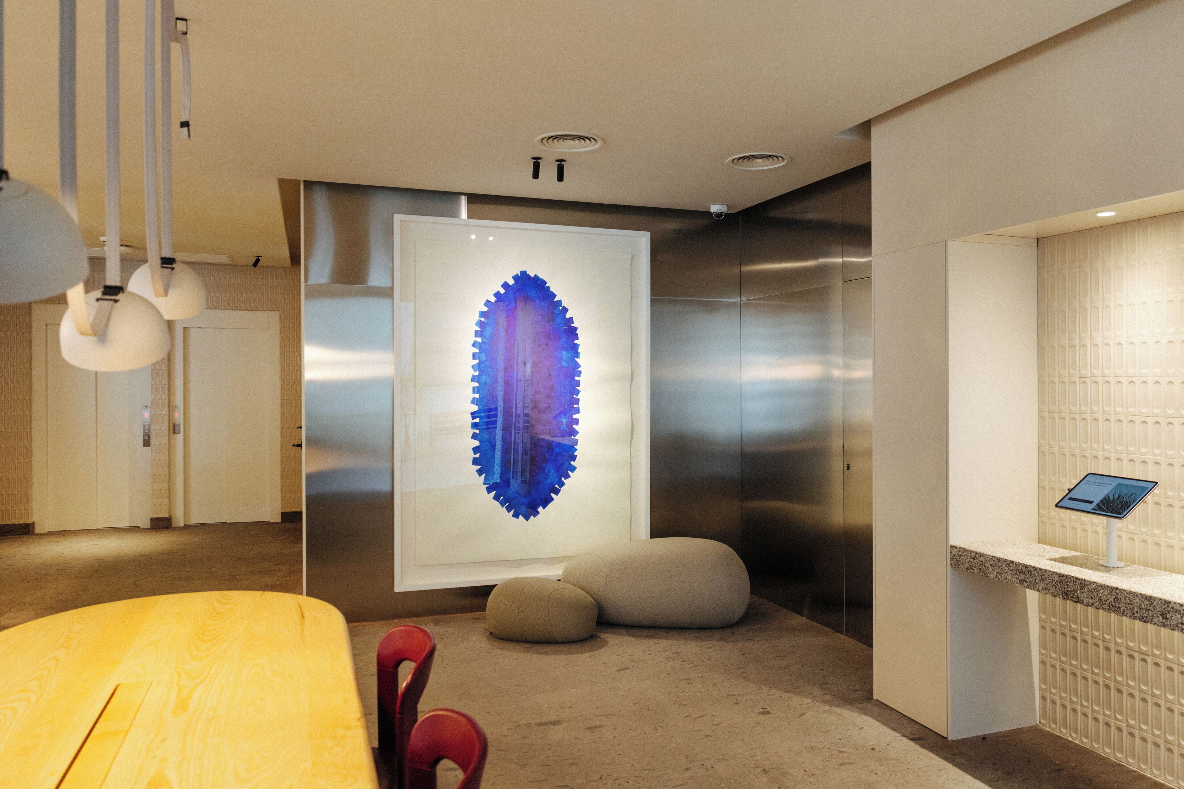 Een modern interieur met een houten tafel, rode stoelen, een groot abstract kunstwerk en een receptiebalie met een digitaal scherm.
