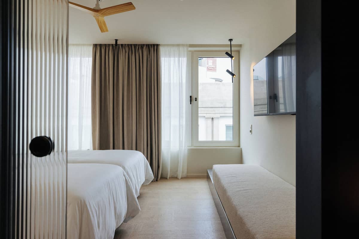 Quarto de hotel com duas camas, ventoinha no teto, janela com cortinas e televisão de tela plana fixada na parede.