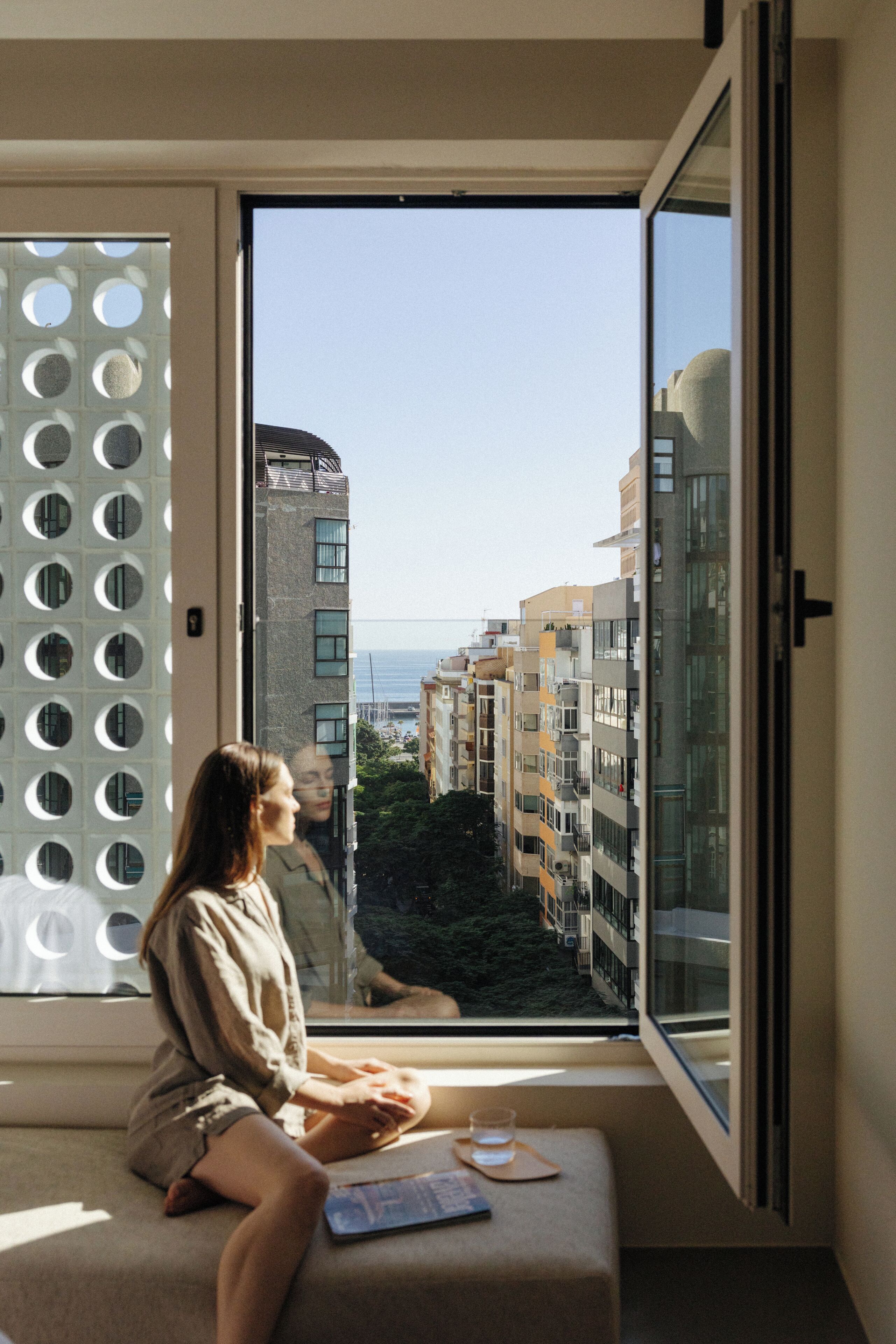 Alguém sentado perto de uma janela, com vista para edifícios e um corpo de água.