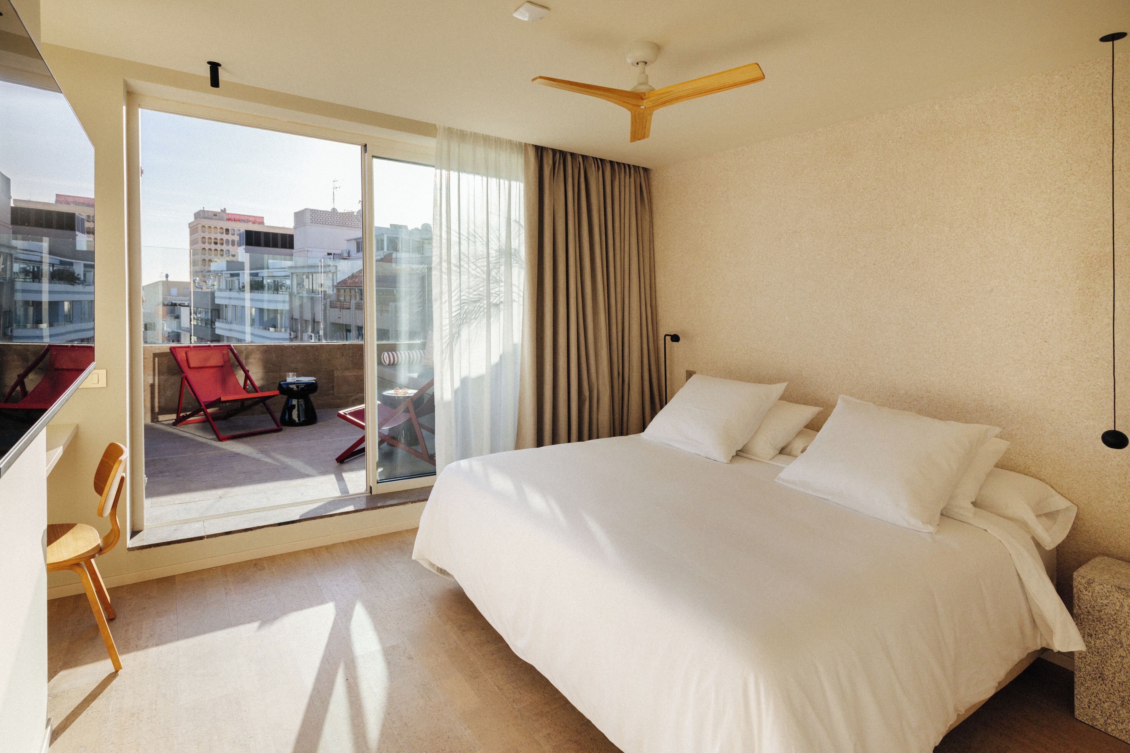 Una habitación de hotel con una cama grande, un balcón con sillas rojas y vistas a la ciudad.