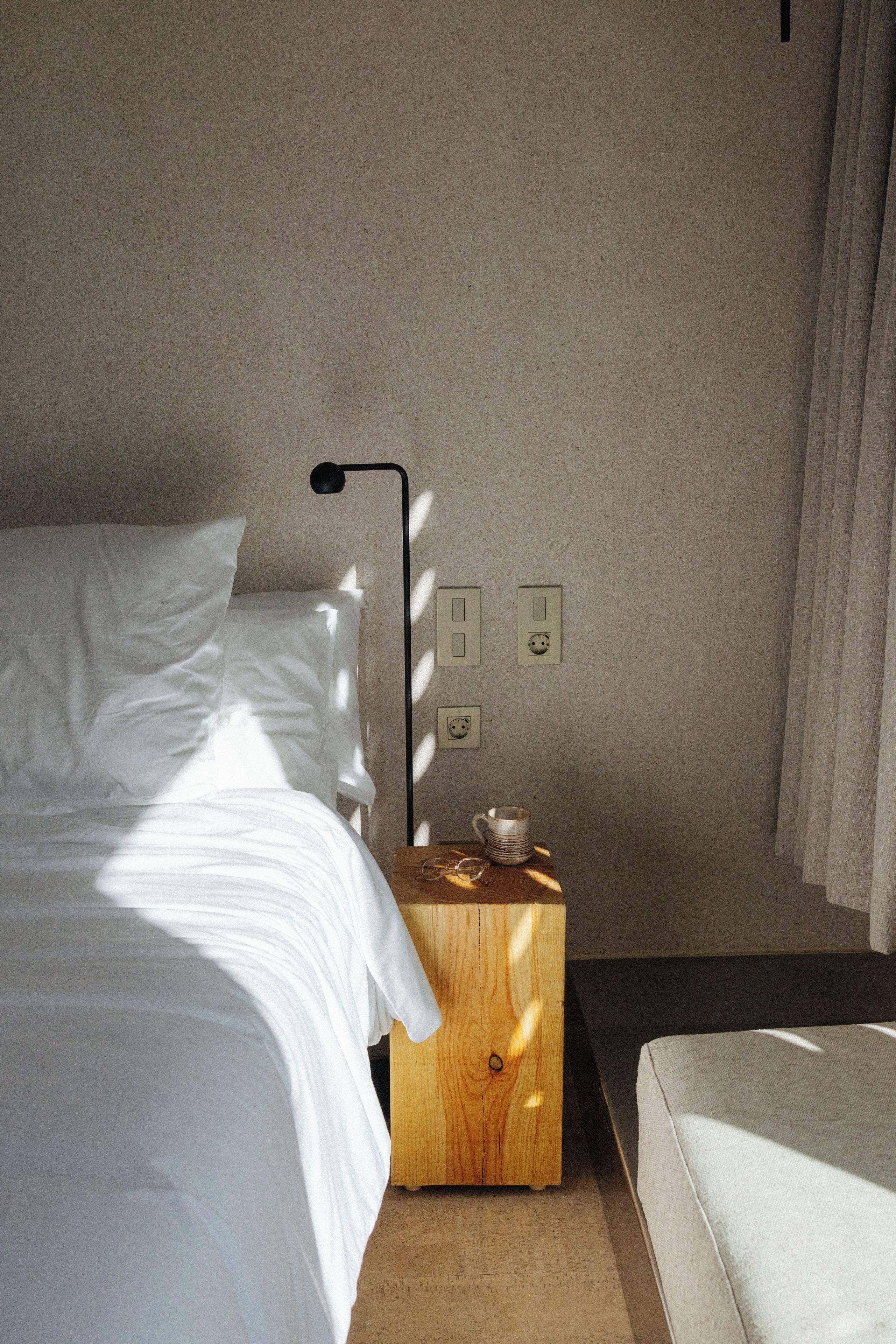 A neatly made bed with white linens, a bedside table with a lamp, and a power outlet on the wall.