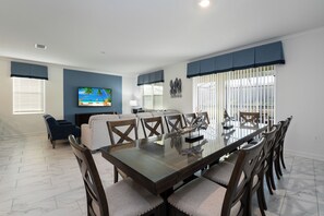 A dining area with a glass-top table and wooden chairs, a sofa, and a TV on the wall. - Awesome 8BR Villa Pool Theater Arcade (Kissimmee)