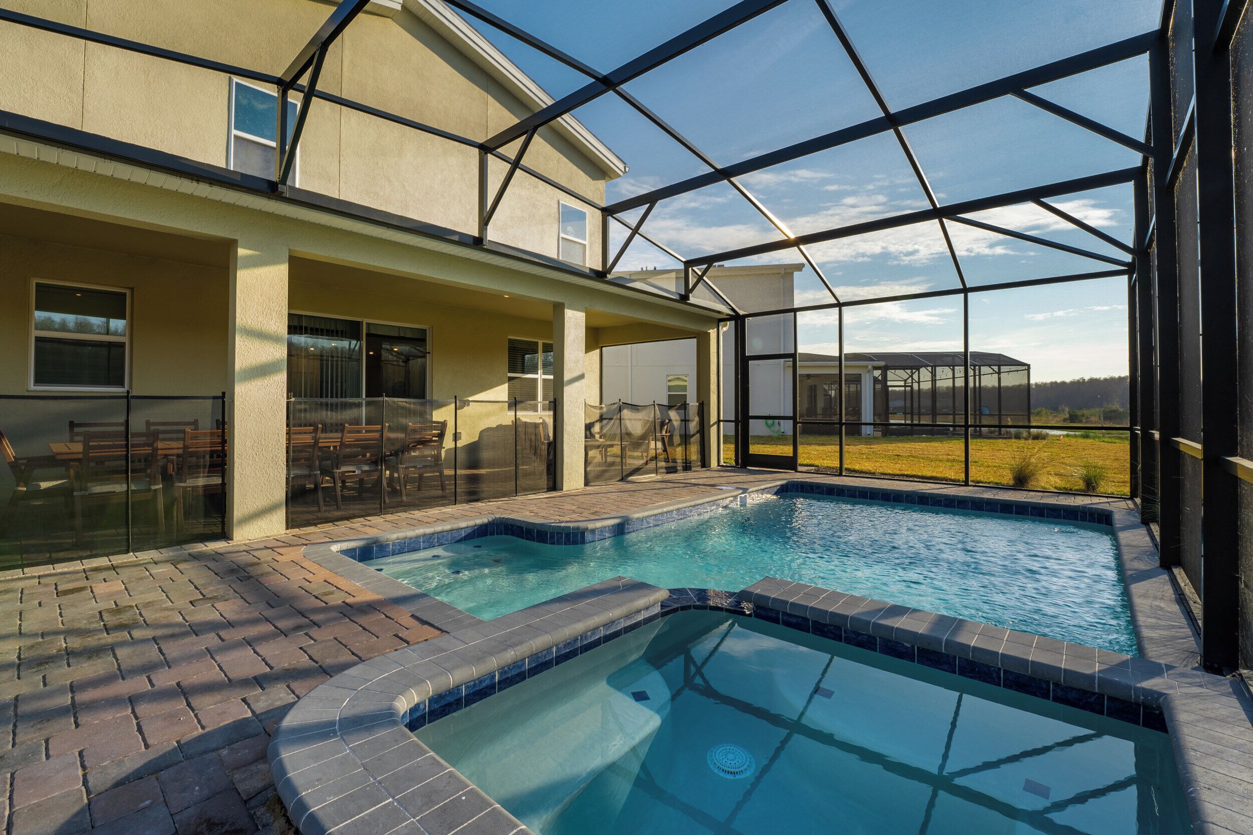 A swimming pool area with a glass enclosure, a patio with outdoor furniture, and a view of a field.