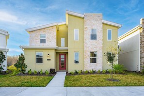 A two-story residential building with a stone facade, a red door, and a small front yard with young trees. - Awesome 8BR Villa Pool Theater Arcade (Kissimmee)
