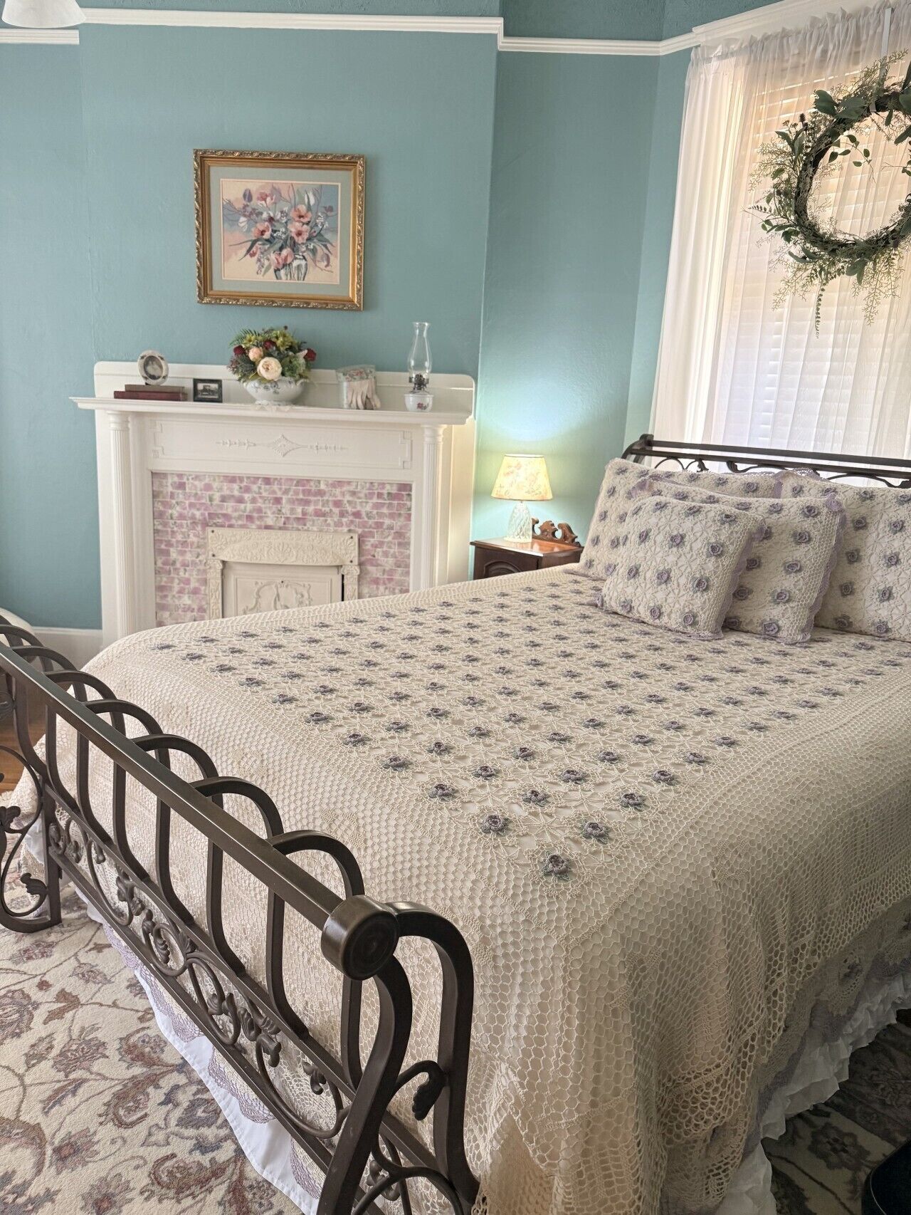 A bedroom with a bed, a fireplace, a wall-mounted picture, and a window with a wreath.