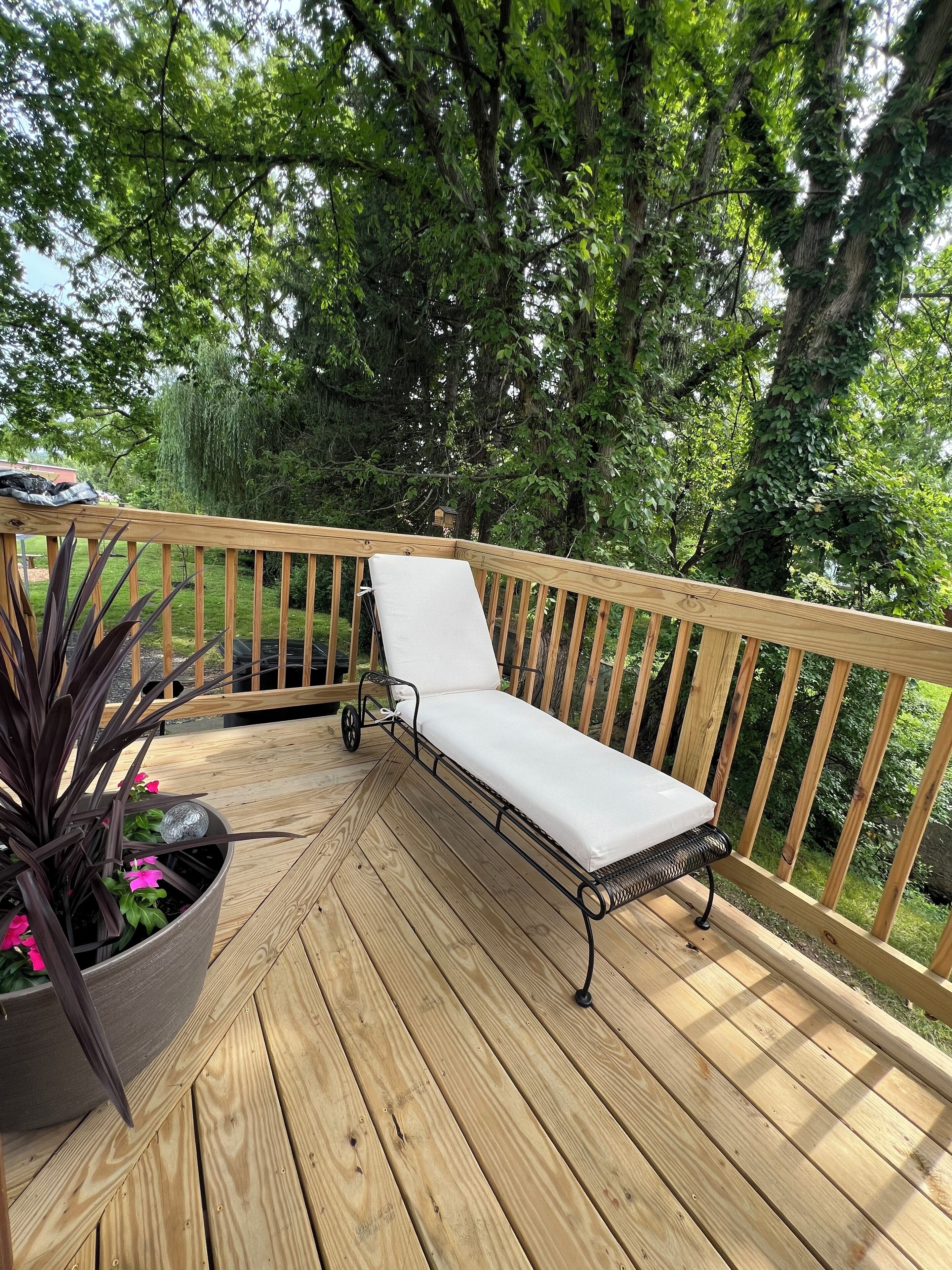 A wooden deck with a metal-framed lounge chair and a potted plant with pink flowers.