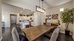 Un comedor espacioso con una mesa de madera y cuatro sillas, una cocina al fondo y una lámpara colgante.