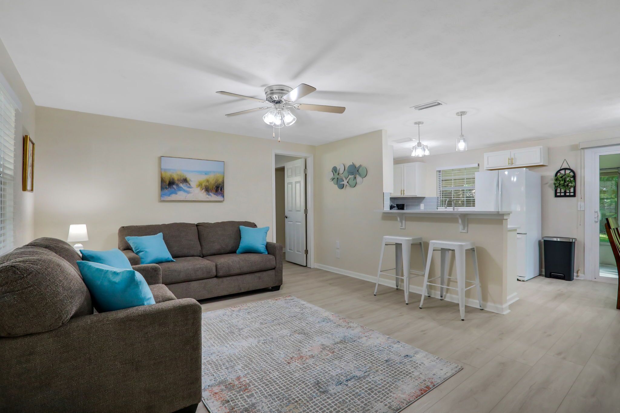A living room with a brown sofa, a ceiling fan, and a kitchen area with white cabinets and a refrigerator.