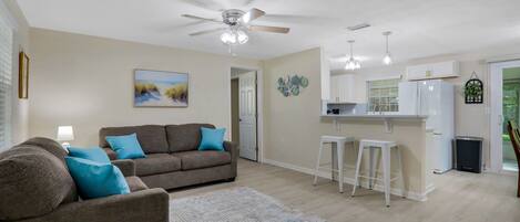A living room with a brown sofa, a ceiling fan, and a kitchen area with white cabinets and a refrigerator.