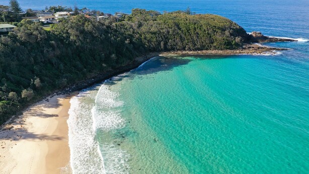 Vue aérienne d’une zone côtière avec une plage de sable, des eaux turquoise et limpides, et une colline boisée.